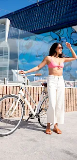 A woman in sunglasses poses by a white bicycle at sunny harbor beside a blue mural wall.