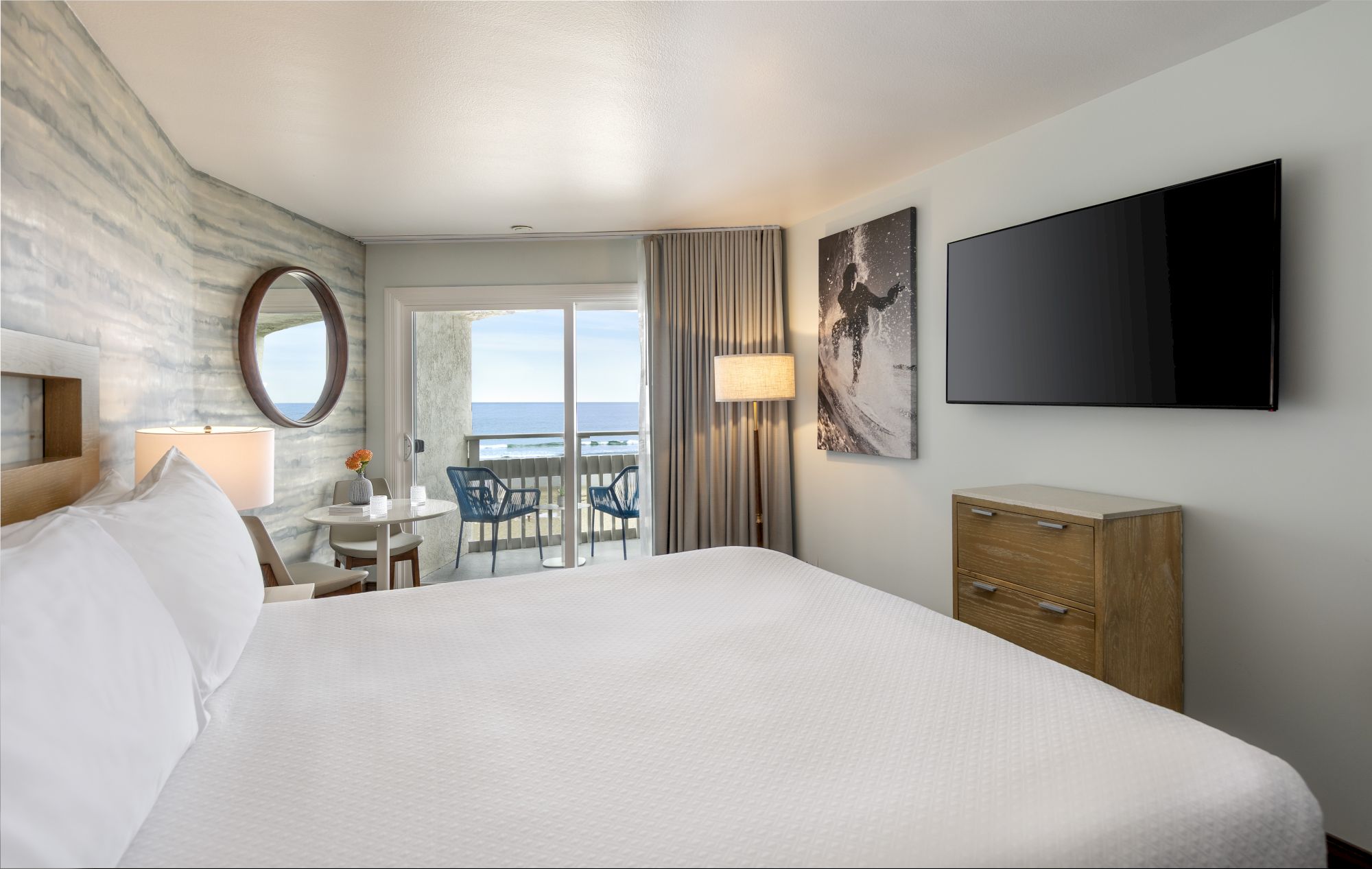 A hotel room with a large bed, wall art, a TV, dresser, a round mirror, and a balcony view of the ocean beyond curtains.