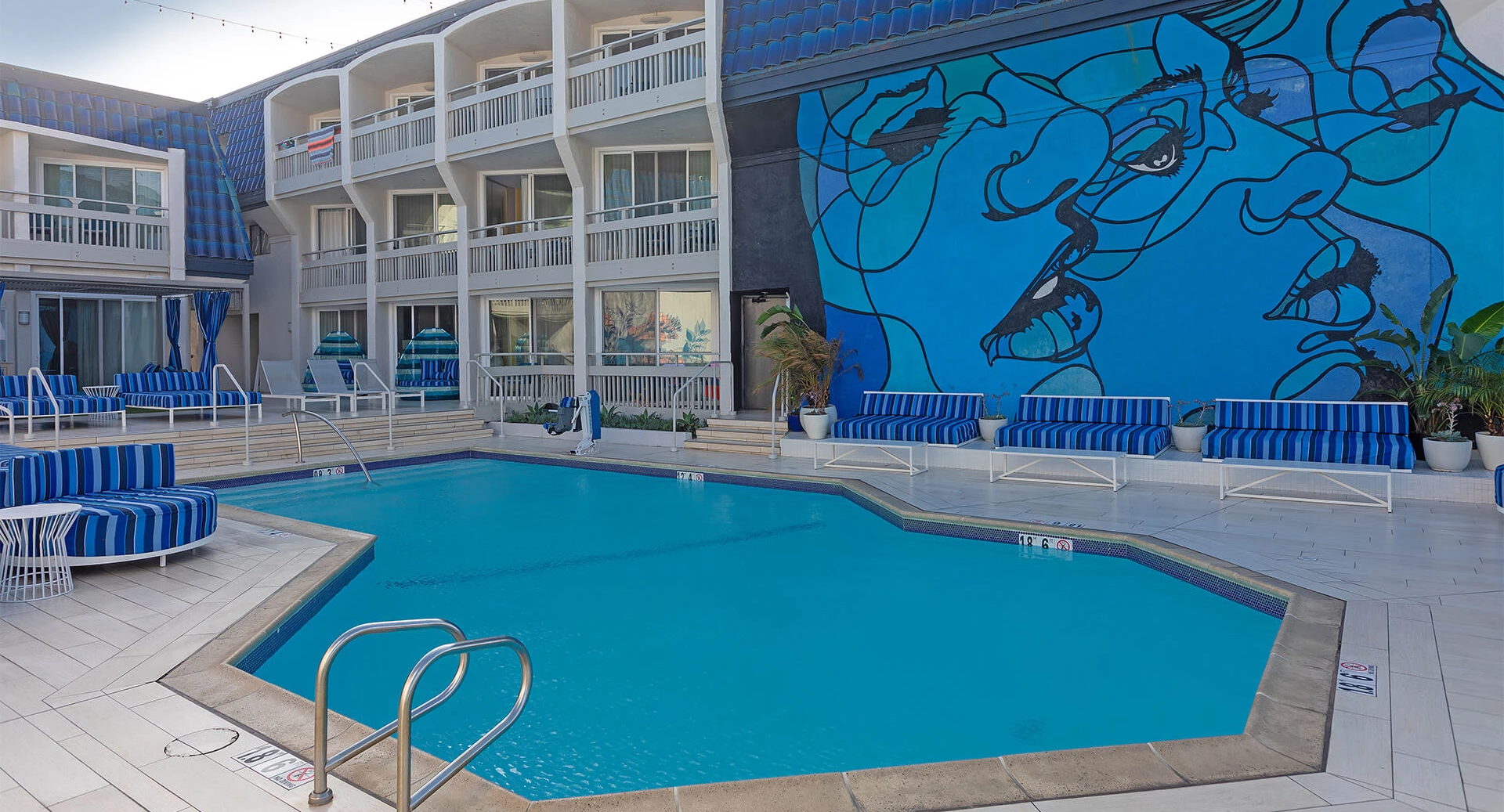 A resort pool area with blue umbrellas, white walkways, and a large blue mural on the building wall featuring abstract faces, relaxing lounge chairs.