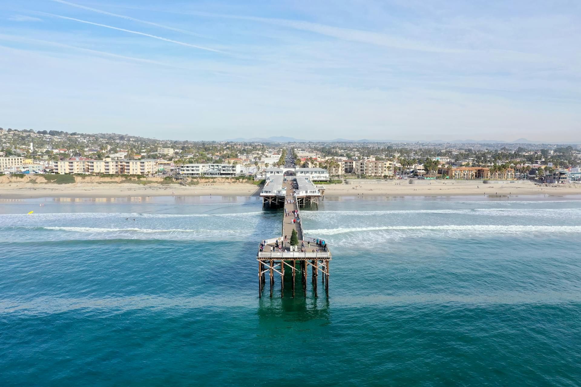 A long pier extends into blue water toward a distant sandy beach with buildings along the shore, under a clear sky.