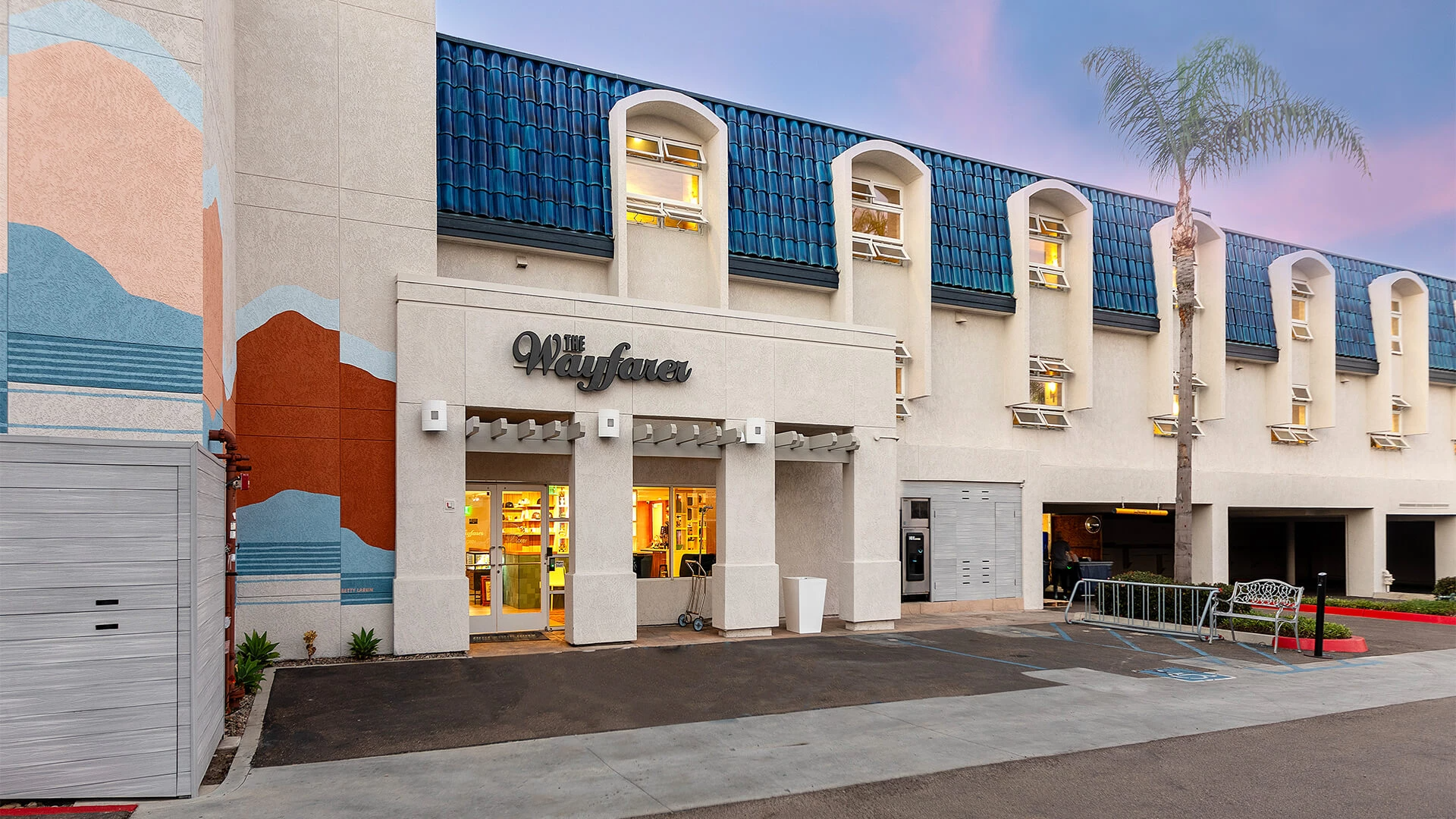 A pastel storefront with curved windows, &ldquo;Wayfair&rdquo; sign, palm trees, and a mural wall; parking and a storefront entrance in a sunny, modern mall facade.