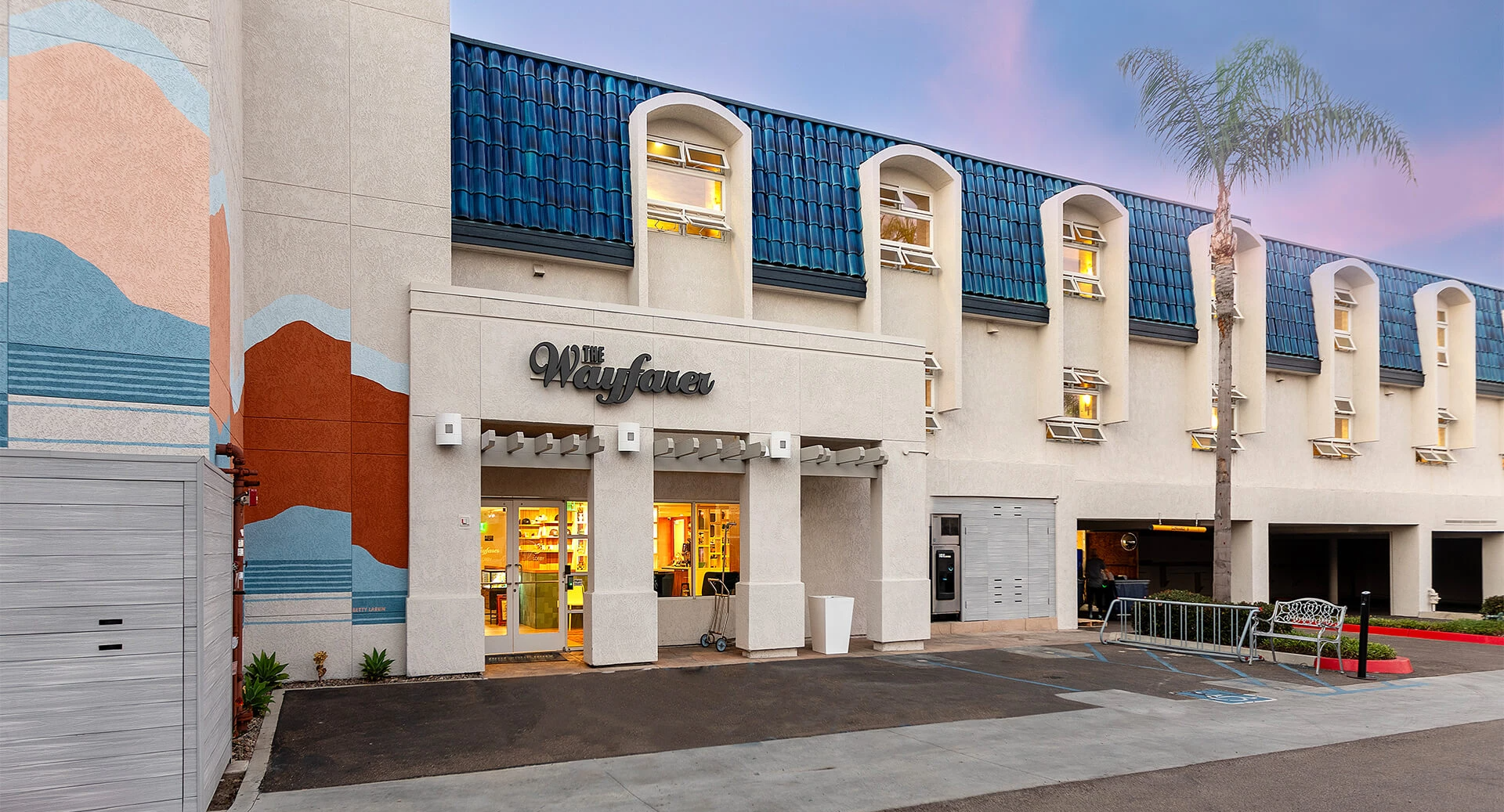A pastel storefront with curved windows, &ldquo;Wayfair&rdquo; sign, palm trees, and a mural wall; parking and a storefront entrance in a sunny, modern mall facade.