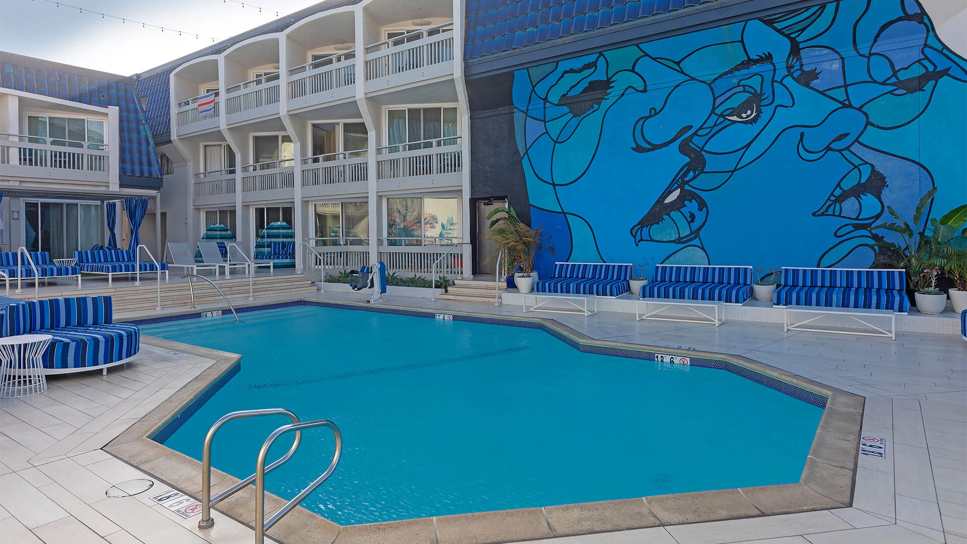 A hotel pool area with blue loungers and a striking blue mural on the wall, surrounded by white balconies and sunny skies. Ending sentence.