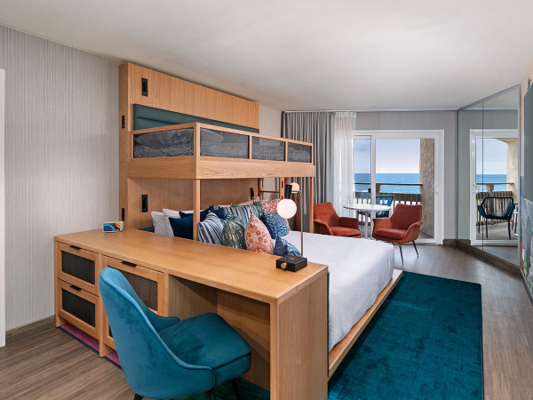 A modern hotel room with a wooden bunk bed, a desk, blue chair, teal rug, and a view of the ocean through large windows in the background.