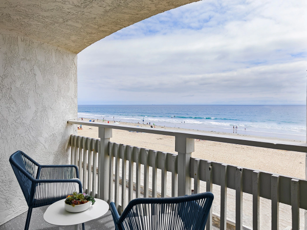 A balcony with two blue chairs and a small table overlooking a sandy beach and blue ocean under a partly cloudy sky.