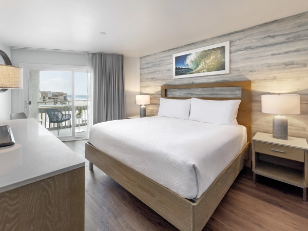 A modern hotel room with a king bed, wood accents, two bedside lamps, a desk, and a balcony with ocean view.