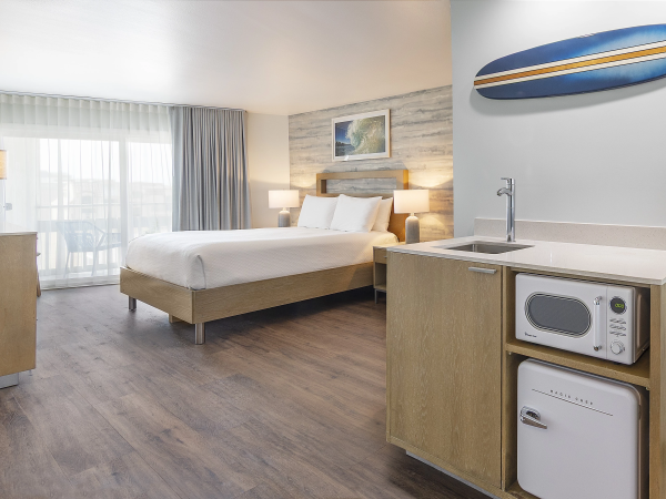 A modern hotel room with a bed, nightstands, a kitchenette (microwave and mini-fridge), wood floors, and a surfboard wall decor, bright and airy.