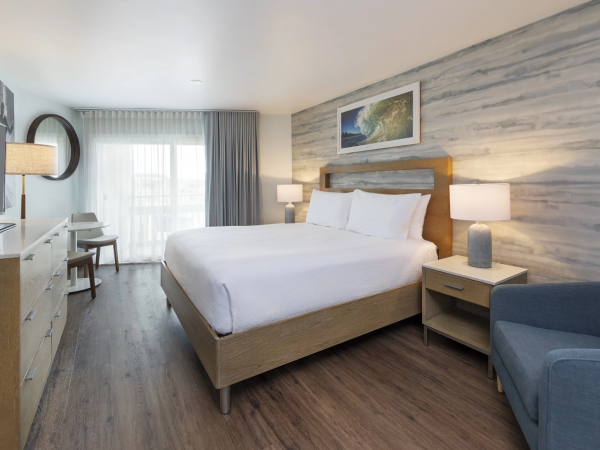 A bright hotel room with a wooden bed, marble-like wall, two nightstands, lamps, a blue sofa, desk, and a large window with sheer curtains, ending this sentence.