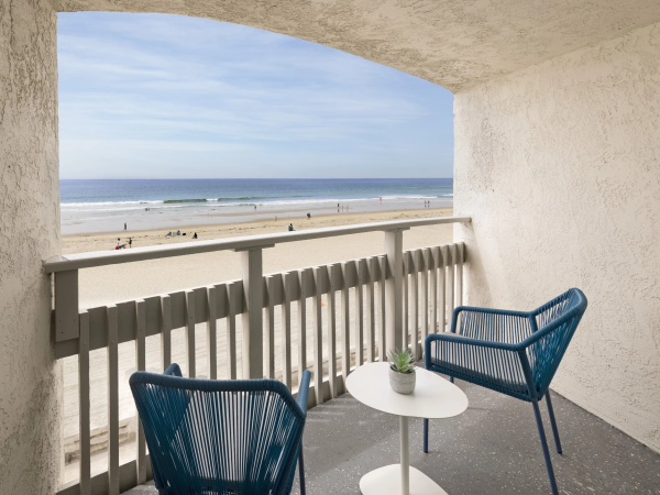 A balcony view of a sunny beach with blue chairs and a small table overlooking the sandy shore and ocean, peaceful and inviting.