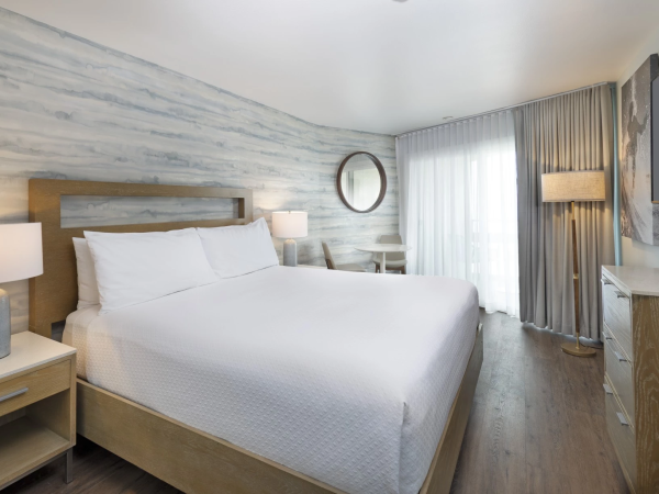 A modern hotel room with a large bed, wooden furniture, soft lighting, a round mirror, and a balcony door with sheer curtains.