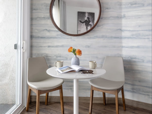 A small round table with two beige chairs, a vase of orange flowers, magazines, and a framed photo reflected in a round wall mirror.