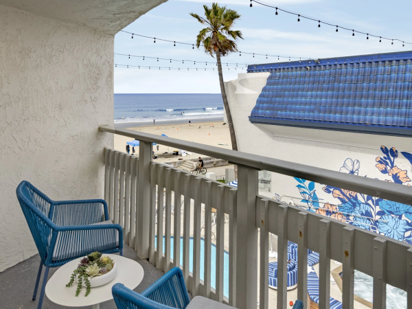 A cozy balcony with blue chairs, a small table, string lights, and a view of the sandy beach and ocean beyond.