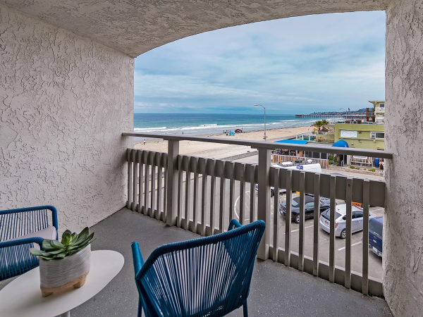 A balcony view over a beach and sea, with white-washed walls, blue chairs, a small table with a plant, and a street below near shops.