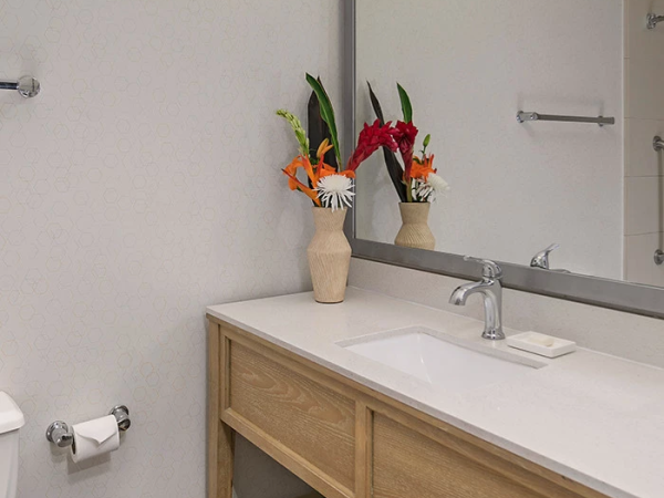 A modern bathroom with a wooden vanity, large mirror, bright light, and fresh flowers in vases on the counter.