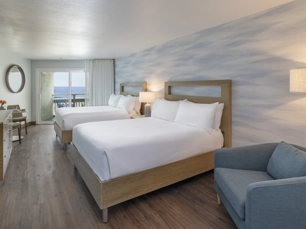 A bright hotel room with two queen beds, a blue upholstered chair, wooden floor, a desk, wall mural of a cloudy sky, and a balcony view to the sea, end.