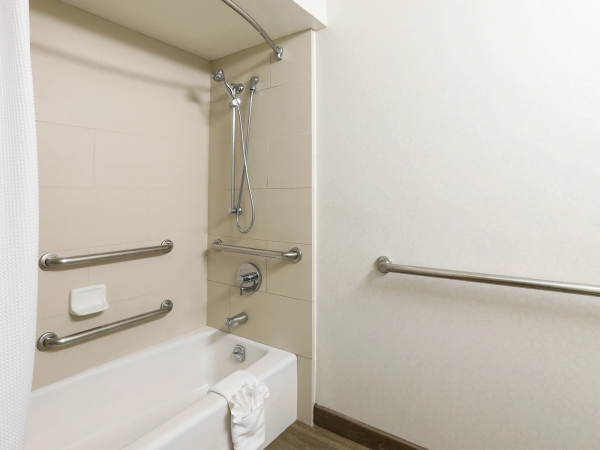 A accessible bathroom with a bathtub/shower combo, grab bars on walls, a handheld shower, a shower curtain, and a small towel on the tub edge, all in a clean, light-colored space.