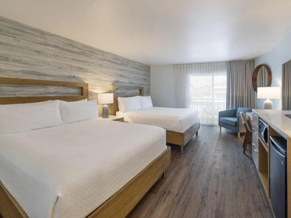 A hotel room with two large beds, a desk, a chair, a TV, and a sliding glass door to a bright balcony, modern decor and wood-style flooring.
