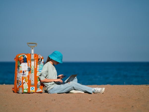 A person in a blue hat works on a laptop on a beach next to an orange, sticker-covered suitcase by the ocean.