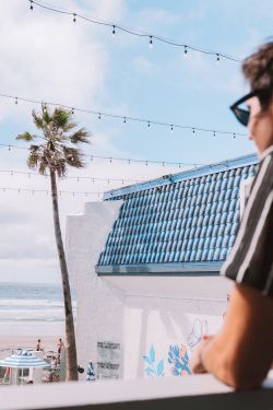 A man in a striped shirt and sunglasses leans on a balcony, overlooking a beach with a palm tree, string lights, and the ocean in the distance.