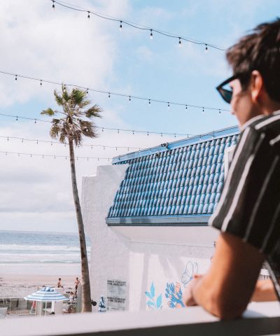 A man in a striped shirt and sunglasses leans on a balcony, overlooking a beach with a palm tree, string lights, and the ocean in the distance.