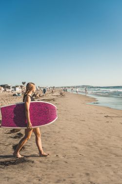 A person walking along the sunny beach carrying a pink surfboard toward the ocean, with other beachgoers in the distance.