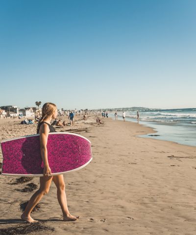 A person walking along the sunny beach carrying a pink surfboard toward the ocean, with other beachgoers in the distance.