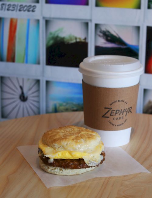 A coffee cup and a breakfast sandwich on a wooden table, with a colorful photo wall in the background, ending the sentence.