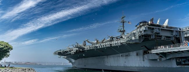 A military ship docked near a rocky coastline under a blue sky, with calm water and a few seagulls nearby.