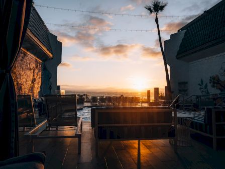 A sunset view at a rooftop lounge with seating, string lights, and a palm tree, casting warm orange glow over chairs and a sea horizon.