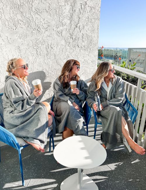 Three women in robes sit on balcony chairs, sipping drinks and relaxing by a white table, with a sunny ocean view behind them.