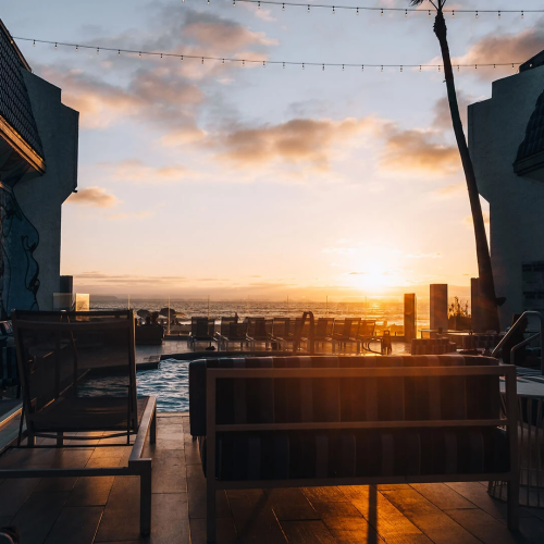 Sunset over a chic rooftop lounge with couches, string lights, and city skyline silhouettes. The warm glow casts long shadows across the seating.
