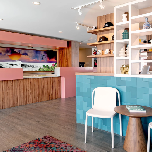 A stylish, coastal-inspired lobby with pink curved counter, wood paneling, turquoise tiled bar, white chairs, and open shelving displaying decor.
