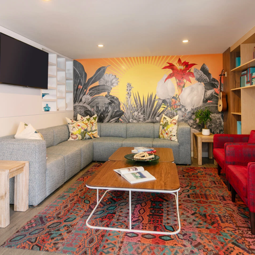 A bright, cozy living room with a red sofa, wooden coffee table, white shelves, and warm lighting; plants and books create a welcoming vibe.