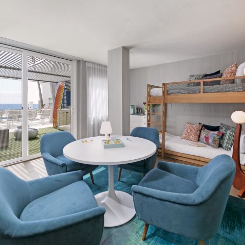 A cozy hotel suite with two queen beds, a large window, a seating area, and a modern kitchenette in warm, inviting tones.