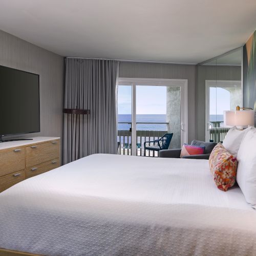 A cozy hotel room with two neatly made beds, soft lighting, a wall-mounted TV, a desk, and a window offering natural light.