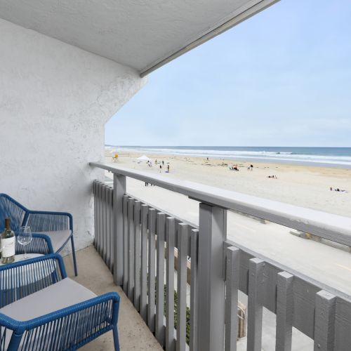 A small balcony with a blue lawn chair, a matching blue seat cover, a side table, and a railing overlooking a bright sky and distant buildings.