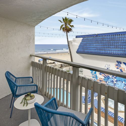 A sunny balcony with blue chairs facing a blue sea and sky, a palm tree in view, and a light railing creating a serene coastal scene.