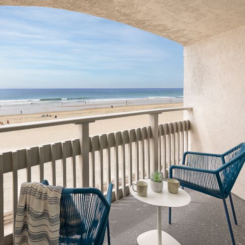 A balcony with a table and two chairs overlooking a coastline; sunny day, calm sea, light-colored walls, railing, and a clear sky.