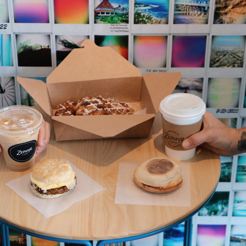A small table with a dozen cupcakes, a few chocolate treats, and a mug, all arranged on a wooden surface with a bright classroom backdrop.