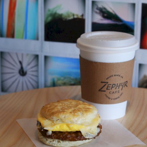 A close-up shows a sausage biscuit on a napkin beside a coffee cup on a wooden table, with a colorful grid mural in the background.
