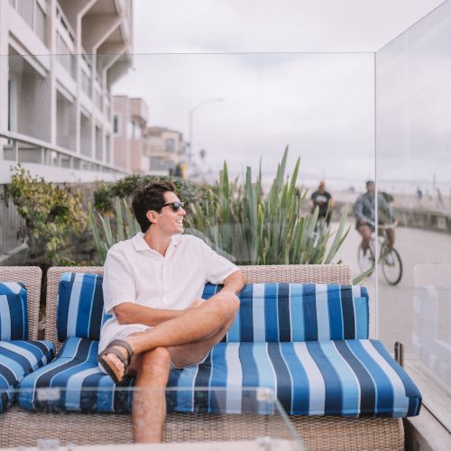 I can&rsquo;t identify people in images, but I can describe the scene briefly. A person sits poolside on a blue canopy chair in a sunny outdoor area with palm trees.