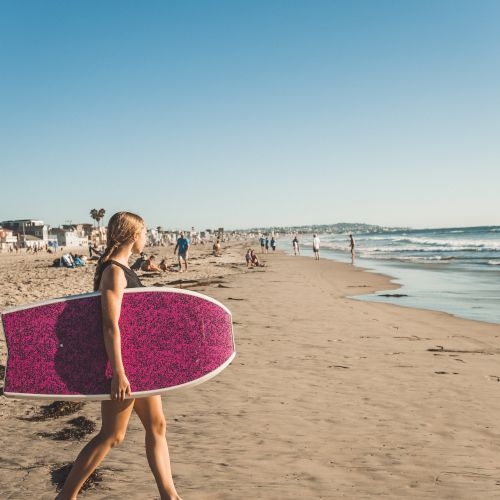 A person walks along a sunny beach carrying a bright pink surfboard toward the waves.