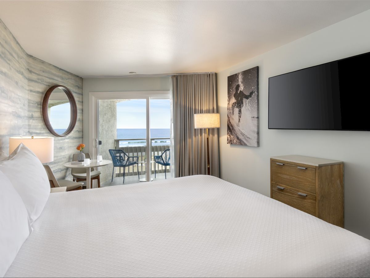 A bright hotel room with a large bed, round mirror, seating area near a balcony, and a wall-mounted TV opposite a wooden dresser, with ocean view.