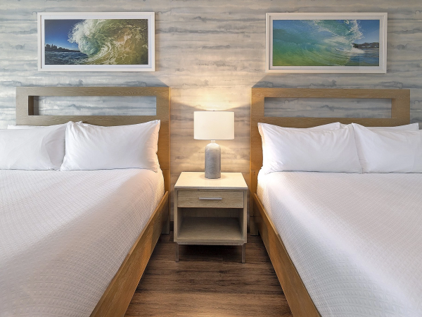 Two twin beds with white linens flank a shared nightstand and lamp, framed by ocean-themed wall art above, in a light wood motel-style room.