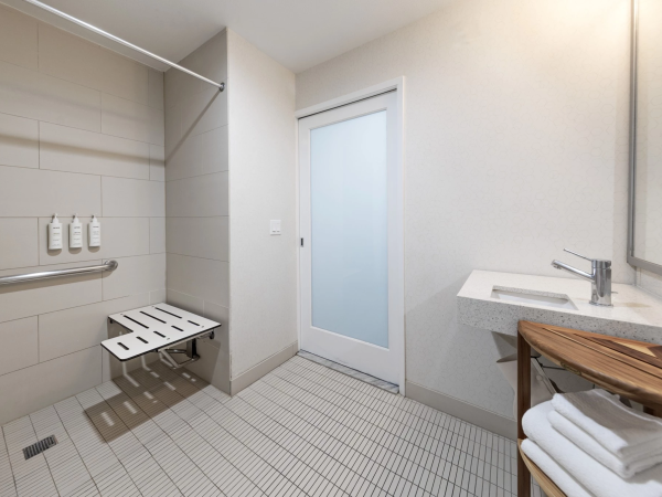A clean, modern bathroom with a wall-mounted bench, beige tiles, a frosted glass door, a single sink, and a towel shelf&mdash;bright and minimalistic.