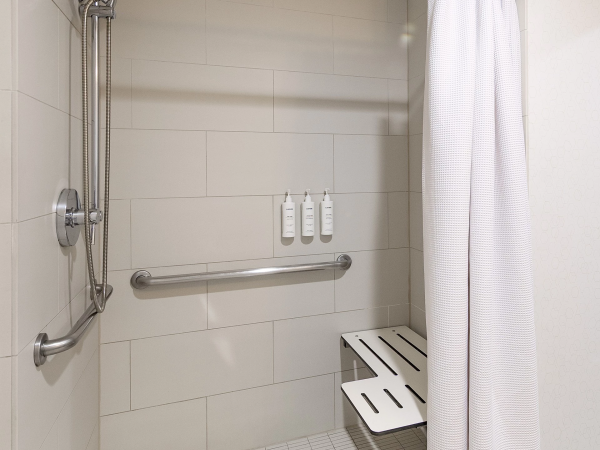 A clean, accessible shower stall with a grab bar, handheld shower, a small bench, and a white curtain in a tiled bathroom.
