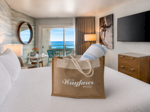 A bright hotel room with a bed, tote bag on the duvet, wooden dresser, wall art, TV, and a balcony showing ocean view.