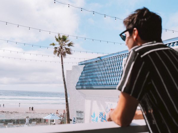 A man in a striped shirt on a balcony overlooking a beach, palm tree, string lights, and a blue-roofed building by the shore, watching the sea.