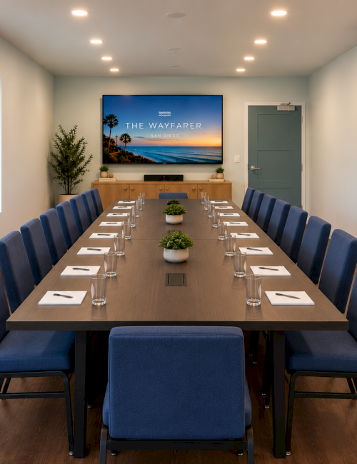 A modern conference room with a long wooden table, blue upholstered chairs, place settings, framed beach photos, and a TV screen.