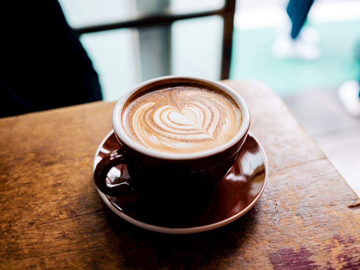 A cup of coffee with latte-art swirl on top sits on a wooden table, with a saucer underneath.